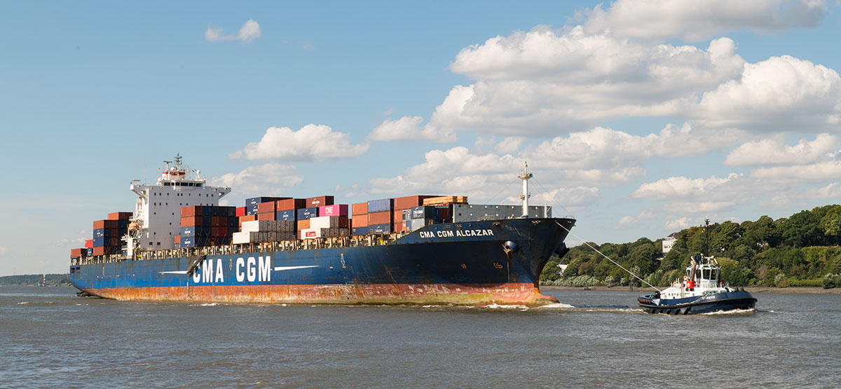 Ein großes mit Containern beladenes Containerschiff wird von einem Schlepper über die Elbe gezogen
